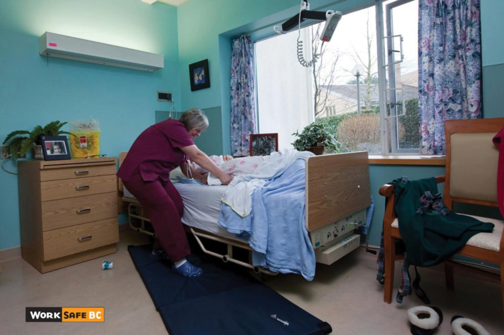 A female healthcare worker in purple scrubs leans over a patient lying in a hospital bed in a brightly lit room with blue walls. A black safety mat is on the floor beside the bed, and a mechanical patient lift is suspended from the ceiling. The room is furnished with a wooden dresser and chair, and there is a large window with patterned curtains. The WorkSafeBC logo is in the bottom left corner.